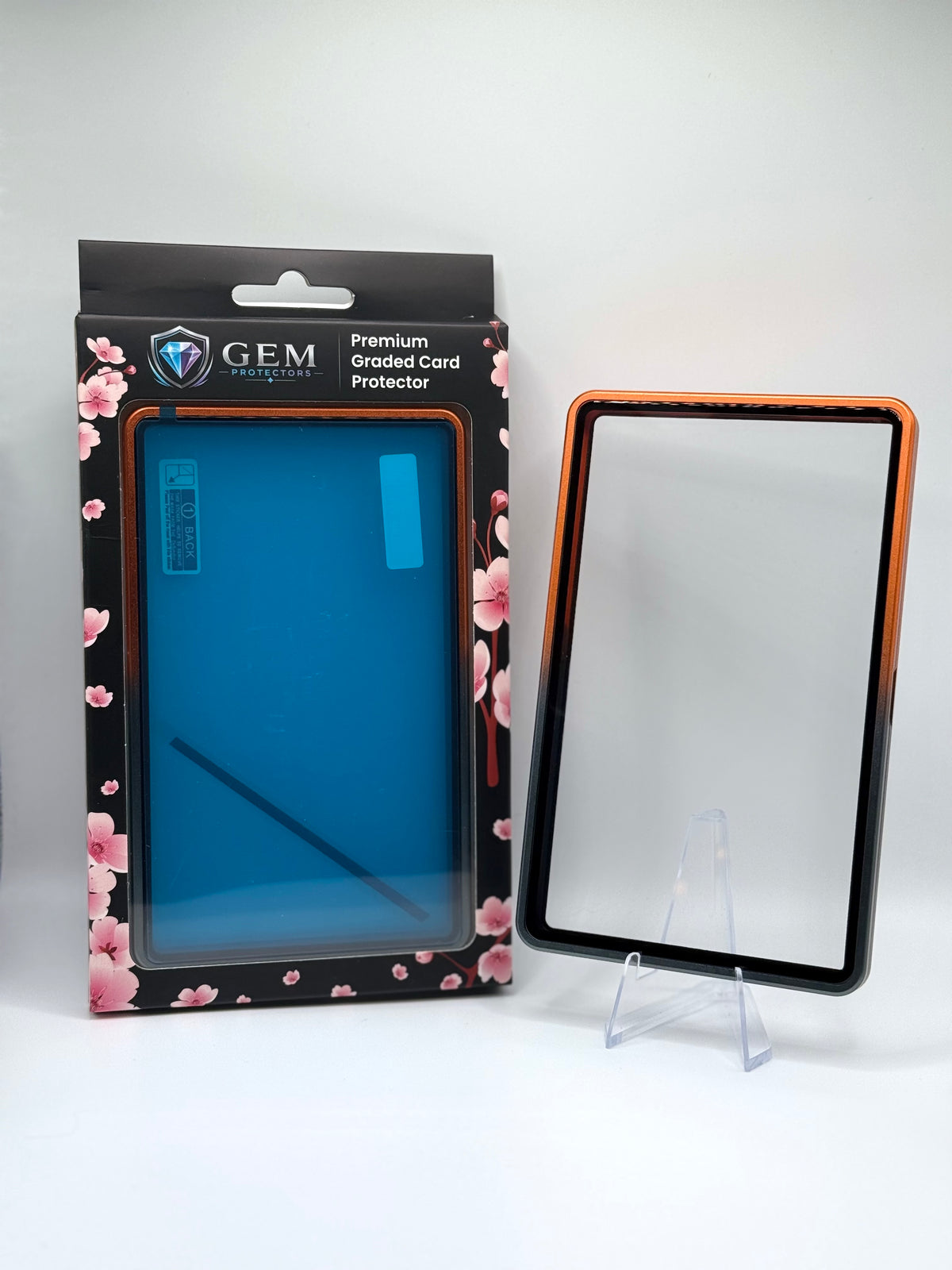 Front view of the empty Black & Orange Gem Protectors case showing the frame and clear panel, beside its retail box